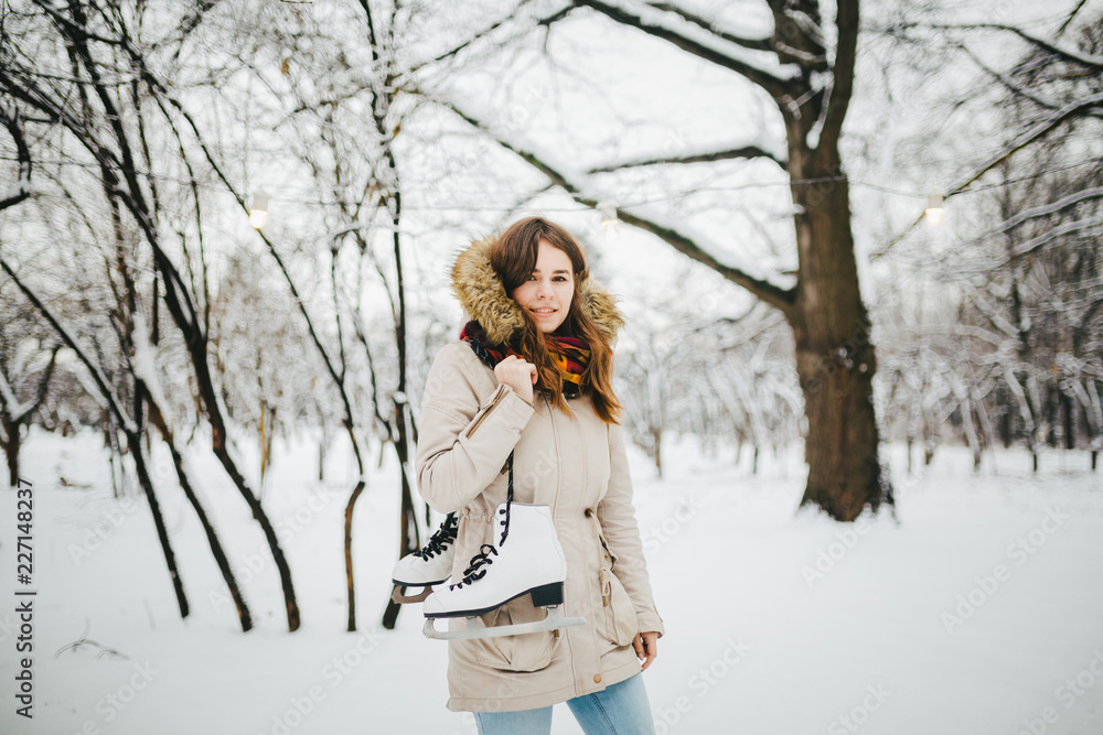 Theme is weekend holiday in winter. A beautiful young Caucasian woman stands in a snow covered park in jacket with hood and fur in jeans and holds on shoulder pair of white skates for long shoelaces Theme is weekend holiday in winter. A beautiful young Caucasian woman stands in a snow covered park in jacket with hood and fur in jeans and holds on shoulder pair of white skates for long shoelaces