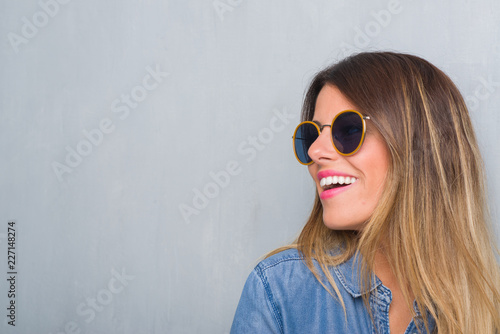 Young adult woman over grunge grey wall wearing retro sunglasses looking away to side with smile on face, natural expression. Laughing confident.