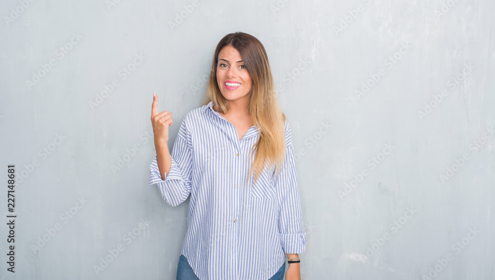 Young adult woman over grey grunge wall wearing fashion business outfit pointing finger up with successful idea. Exited and happy. Number one. Young adult woman over grey grunge wall wearing fashion business outfit pointing finger up with successful idea. Exited and happy. Number one.