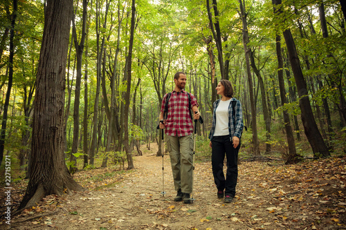 Loving couple wearing checkered shirts hiking side by side through forest on a beautiful autumn day, looking at each other. Healthy and active outdoor lifestyle