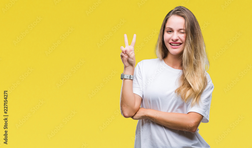 Young beautiful blonde woman wearing casual white t-shirt over isolated background smiling with happy face winking at the camera doing victory sign. Number two. Young beautiful blonde woman wearing casual white t-shirt over isolated background smiling with happy face winking at the camera doing victory sign. Number two.