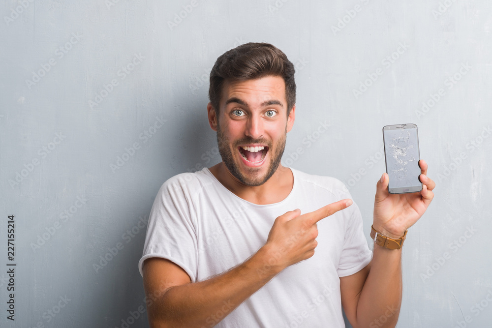 Handsome young man over grey grunge wall showing broken smartphone screen very happy pointing with hand and finger Handsome young man over grey grunge wall showing broken smartphone screen very happy pointing with hand and finger
