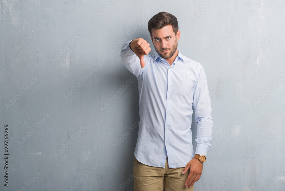 Handsome young business man over grey grunge wall wearing elegant shirt looking unhappy and angry showing rejection and negative with thumbs down gesture. Bad expression.