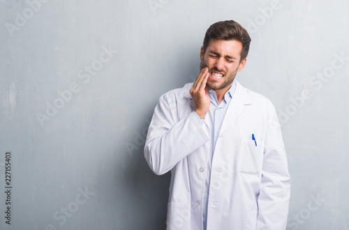 Handsome young professional man over grey grunge wall wearing white coat touching mouth with hand with painful expression because of toothache or dental illness on teeth. Dentist concept.