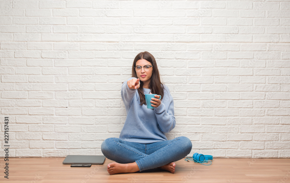 Young brunette woman sitting on the floor over white brick wall drinking coffee pointing with finger to the camera and to you, hand sign, positive and confident gesture from the front Young brunette woman sitting on the floor over white brick wall drinking coffee pointing with finger to the camera and to you, hand sign, positive and confident gesture from the front