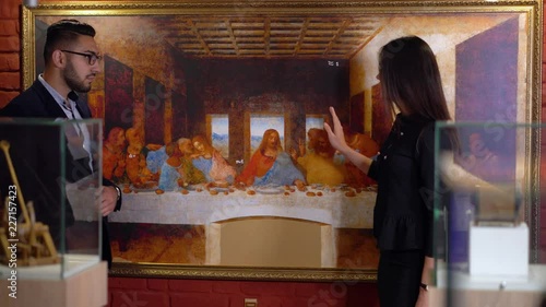 Brunette girl guide tells to the young man in glasses about the features of the exhibition of the famous painting work by Leonardo Da Vinci The Last Supper