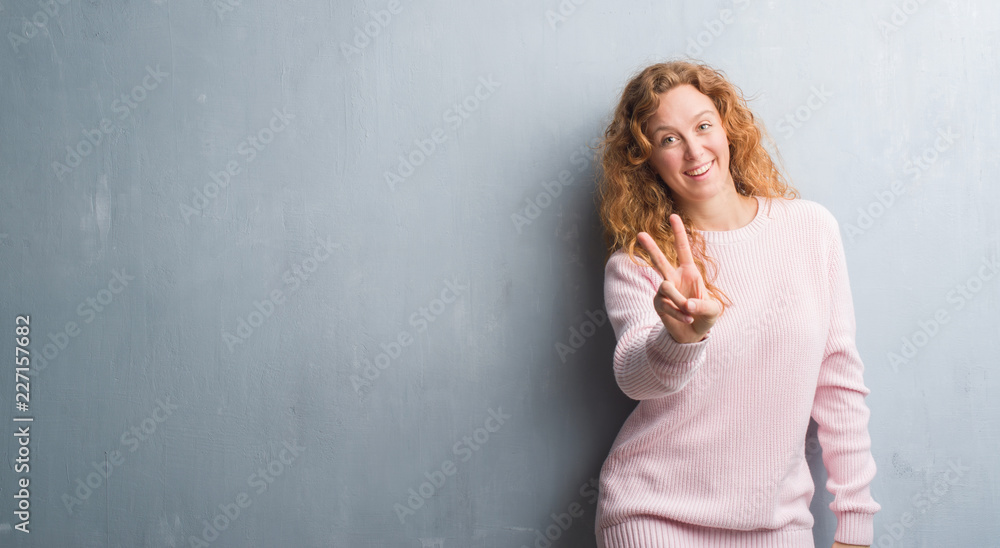 Young redhead woman over grey grunge wall wearing pink sweater smiling looking to the camera showing fingers doing victory sign. Number two. Young redhead woman over grey grunge wall wearing pink sweater smiling looking to the camera showing fingers doing victory sign. Number two.
