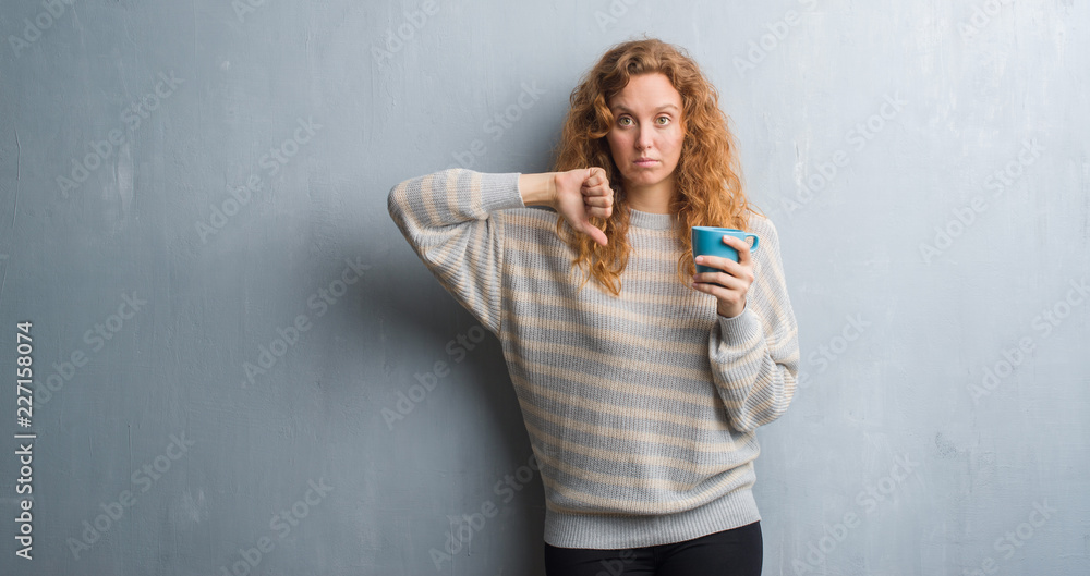 Young redhead woman over grey grunge wall drinking a cup of coffee with angry face, negative sign showing dislike with thumbs down, rejection concept