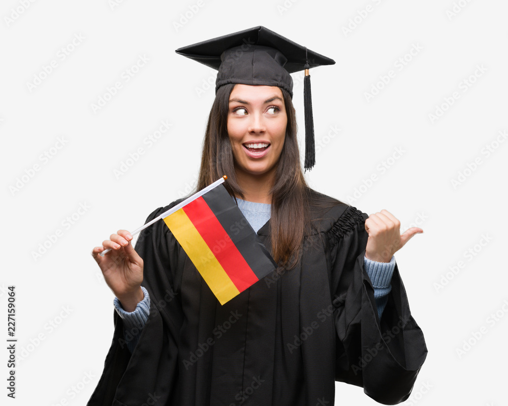 Young hispanic woman wearing graduated uniform holding flag of germany pointing and showing with thumb up to the side with happy face smiling Young hispanic woman wearing graduated uniform holding flag of germany pointing and showing with thumb up to the side with happy face smiling