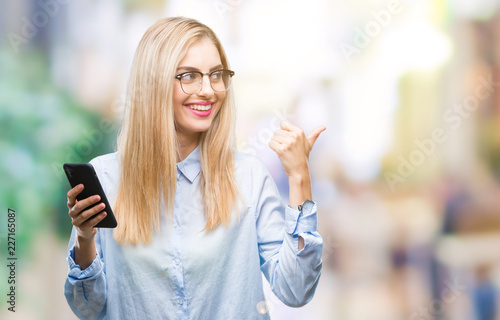 Young beautiful blonde business woman using smartphone over isolated background pointing and showing with thumb up to the side with happy face smiling