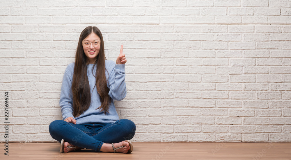 Young Chinese woman sitting on the floor over brick wall showing and pointing up with finger number one while smiling confident and happy. Young Chinese woman sitting on the floor over brick wall showing and pointing up with finger number one while smiling confident and happy.
