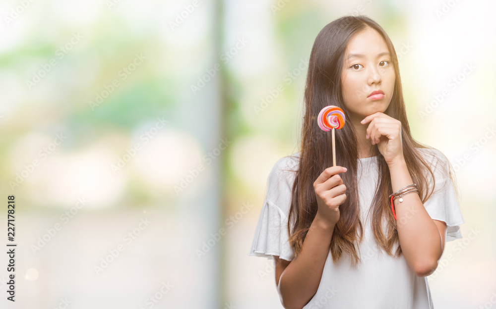 Young asian woman eating lollipop candy over isolated background serious face thinking about question, very confused idea