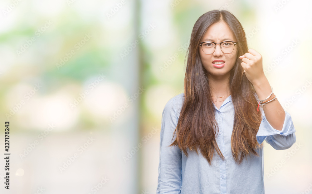 Young asian business woman wearing glasses over isolated background angry and mad raising fist frustrated and furious while shouting with anger. Rage and aggressive concept.
