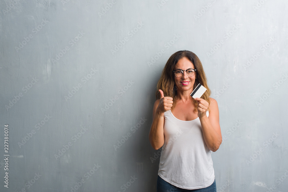 Middle age hispanic woman standing over grey grunge wall holding credit card happy with big smile doing ok sign, thumb up with fingers, excellent sign Middle age hispanic woman standing over grey grunge wall holding credit card happy with big smile doing ok sign, thumb up with fingers, excellent sign