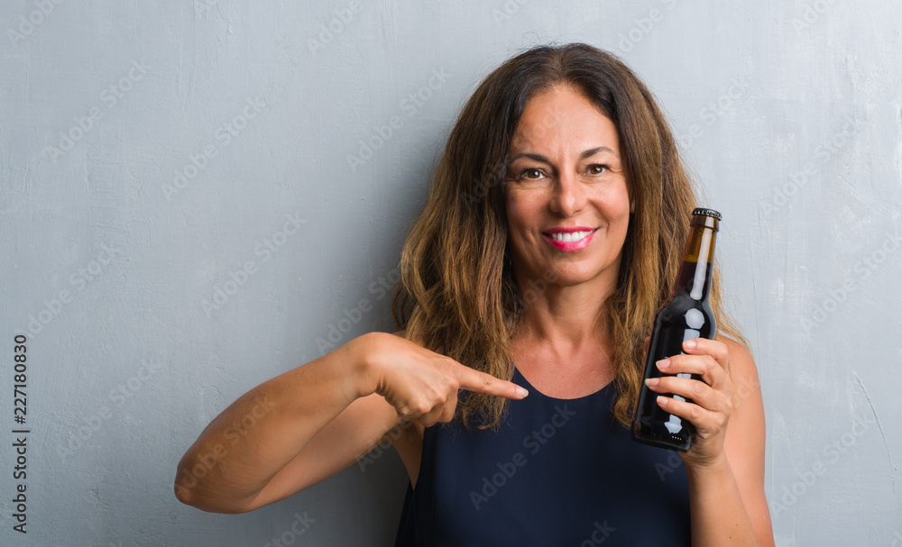 Middle age hispanic woman standing over grey grunge wall holding beer bottle with surprise face pointing finger to himself Middle age hispanic woman standing over grey grunge wall holding beer bottle with surprise face pointing finger to himself