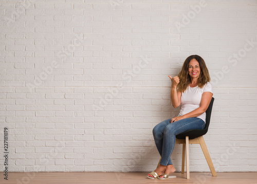 Middle age hispanic woman sitting on chair over white brick walll pointing and showing with thumb up to the side with happy face smiling