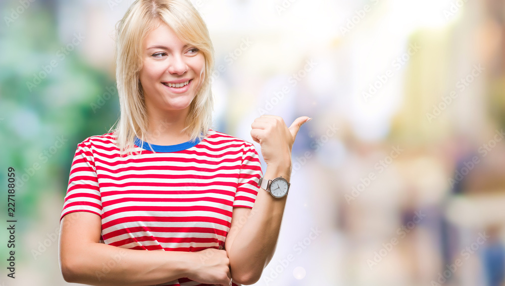 Young beautiful blonde woman over isolated background smiling with happy face looking and pointing to the side with thumb up.