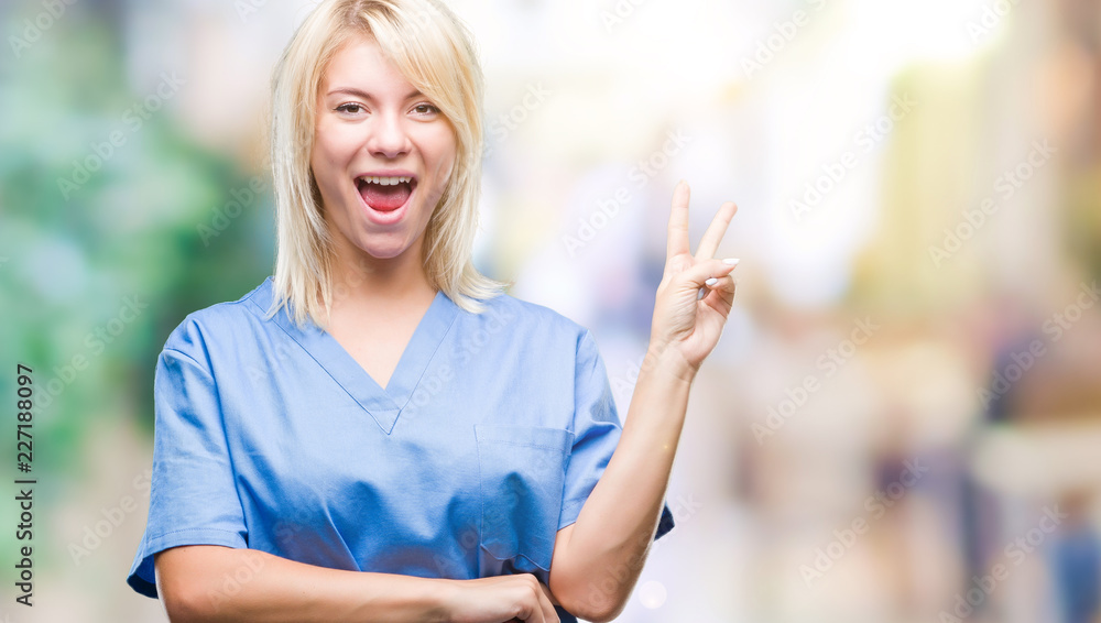 Young beautiful blonde nurse doctor woman over isolated background smiling with happy face winking at the camera doing victory sign. Number two.