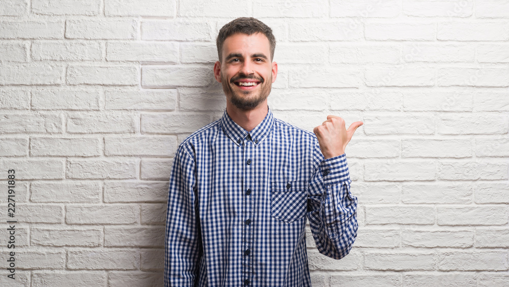 Young adult man standing over white brick wall pointing and showing with thumb up to the side with happy face smiling Young adult man standing over white brick wall pointing and showing with thumb up to the side with happy face smiling