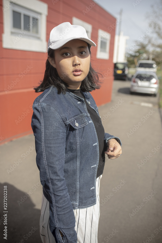 portrait of asian teenager wearing white cap and blue jeans shirt standing outdoor looking to camera