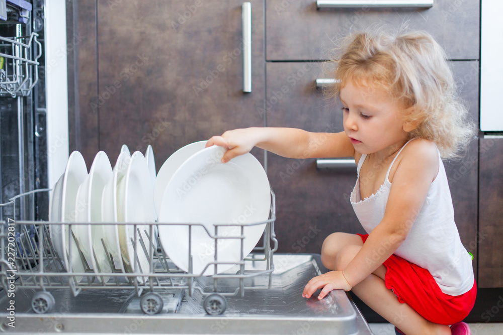 Cute blonde toddler girl helping in the kitchen taking plates out of dish washing machine Cute blonde toddler girl helping in the kitchen taking plates out of dish washing machine