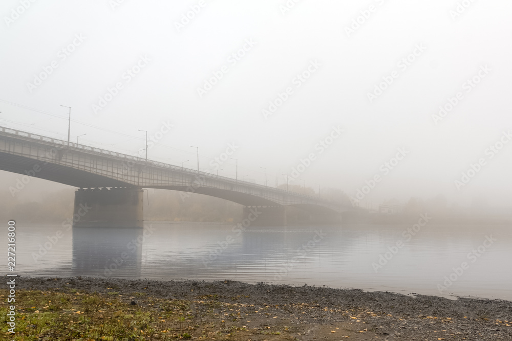 Automobile big bridge stretched across a calm river, wrapped in a dense autumn gray fog.