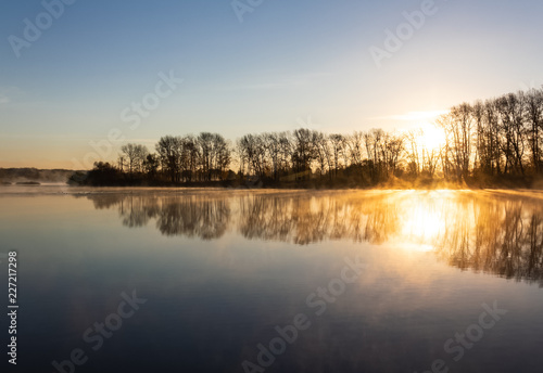 Autumn forest is reflected in the mirror of the river, covered with remnants of fog, which is scattered under the rays of the rising sun.