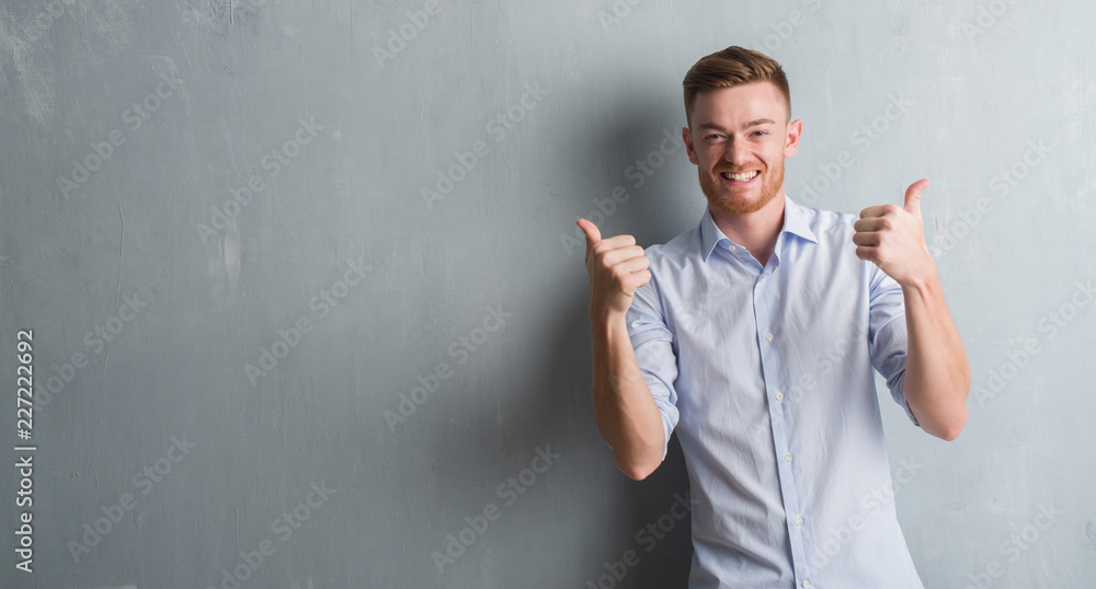 Young redhead business man over grey grunge wall success sign doing positive gesture with hand, thumbs up smiling and happy. Looking at the camera with cheerful expression, winner gesture. - obrazy, fototapety, plakaty 