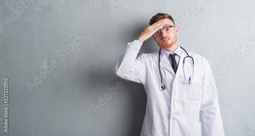 Young redhead doctor man over grey grunge wall stressed with hand on head, shocked with shame and surprise face, angry and frustrated. Fear and upset for mistake.