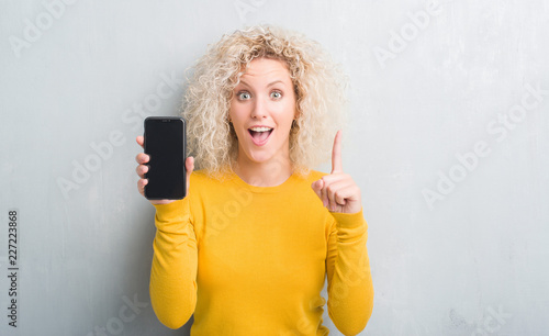 Young blonde woman over grunge grey background showing blank screen of smartphone surprised with an idea or question pointing finger with happy face  number one
