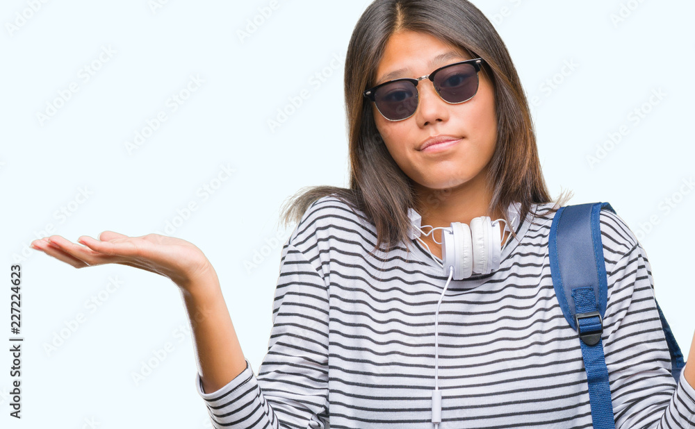 Young asian student woman wearing headphones and backpack over isolated background clueless and confused expression with arms and hands raised. Doubt concept. Young asian student woman wearing headphones and backpack over isolated background clueless and confused expression with arms and hands raised. Doubt concept.