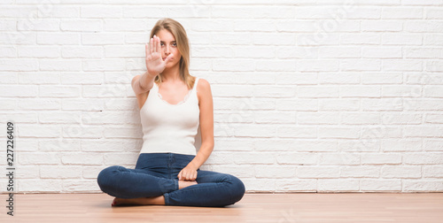 Beautiful young woman sitting on the floor at home doing stop sing with palm of the hand. Warning expression with negative and serious gesture on the face.