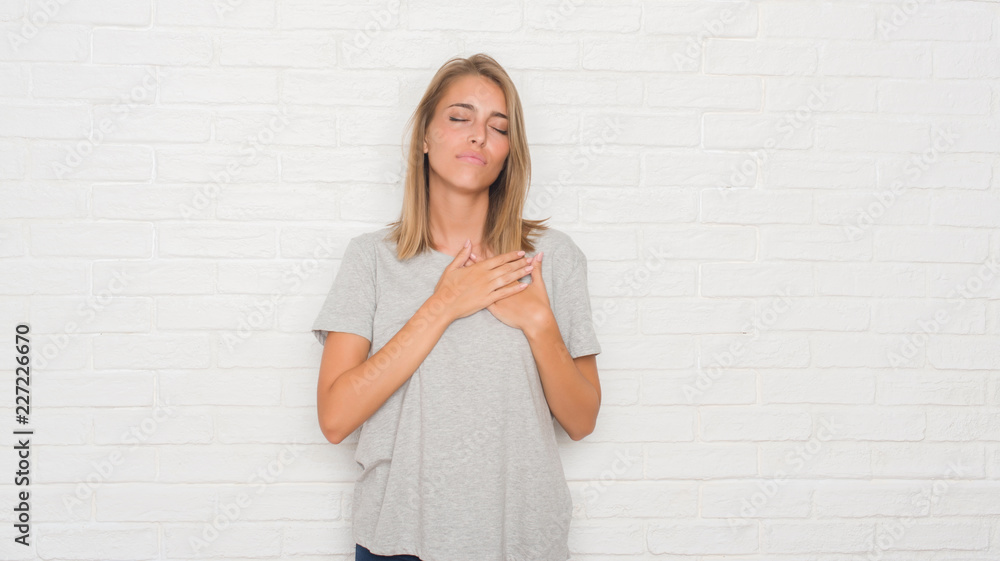 Beautiful young woman over white brick wall smiling with hands on chest with closed eyes and grateful gesture on face. Health concept. Beautiful young woman over white brick wall smiling with hands on chest with closed eyes and grateful gesture on face. Health concept.