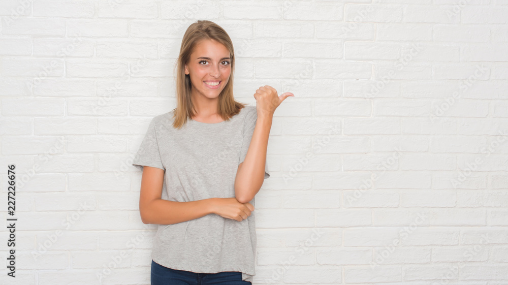Beautiful young woman over white brick wall smiling with happy face looking and pointing to the side with thumb up.