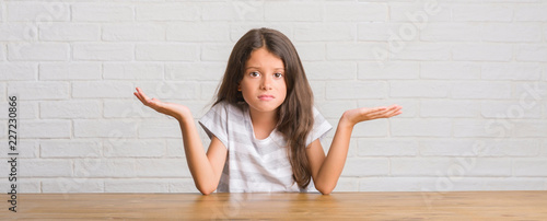 Quadro su tela Young hispanic kid sitting on the table at home clueless and confused expression with arms and hands raised