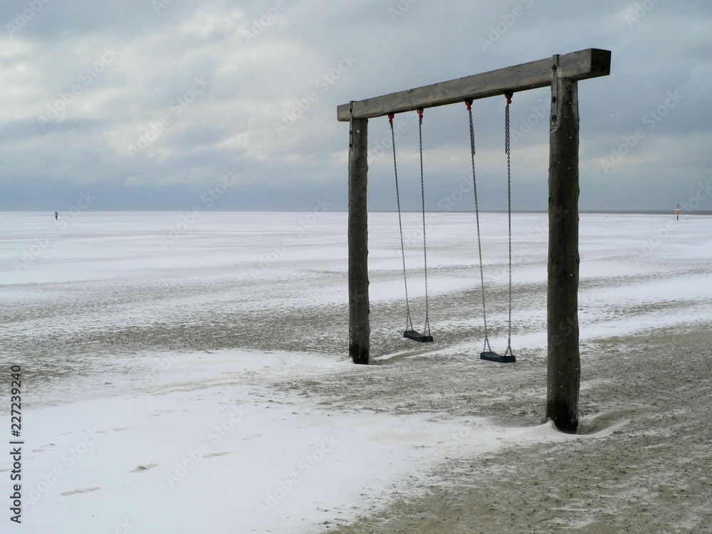 Cold snap - Two swing hang on a wooden beam in the middle of the lonely beach in winter. The sand is mixed with snow and there are some drifts. The weather is cloudy and windy.