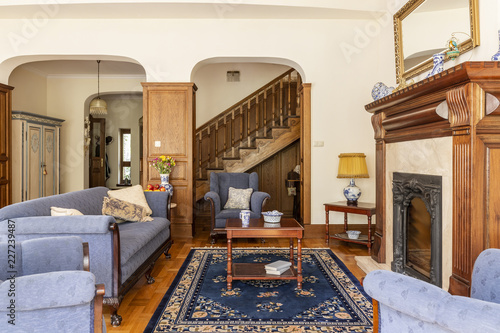 A luxurious living room interior with antique sofa and coffee table in front of a fireplace in an English style mansion. Wooden staircase in the background. Real photo.