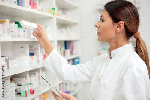 Beautiful young female pharmacist reaching for medications on the shelf, holding a tablet in one hand photo