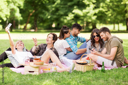 friendship, leisure, technology and people concept - group of friends with smartphones and non alcoholic beer chilling on picnic blanket at summer park