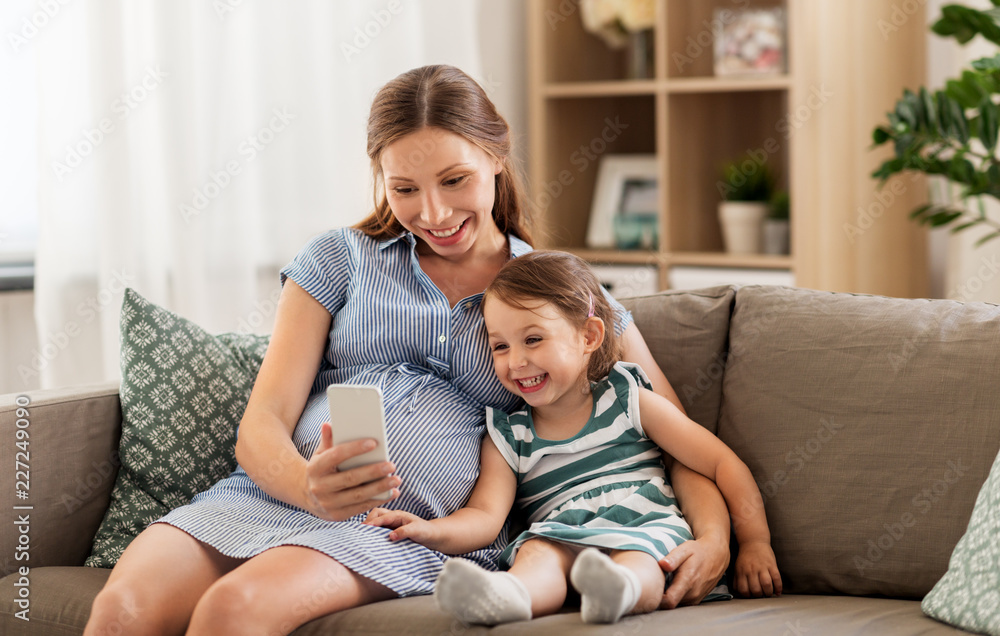 pregnancy, people and family concept - happy pregnant mother and little daughter with smartphone at home