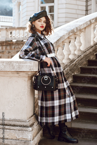 Outdoor full body fashion portrait of young beautiful fashionable girl wearing autumn long checked dress, leather beret, boots, holding black bag, posing in european city, near old architecture