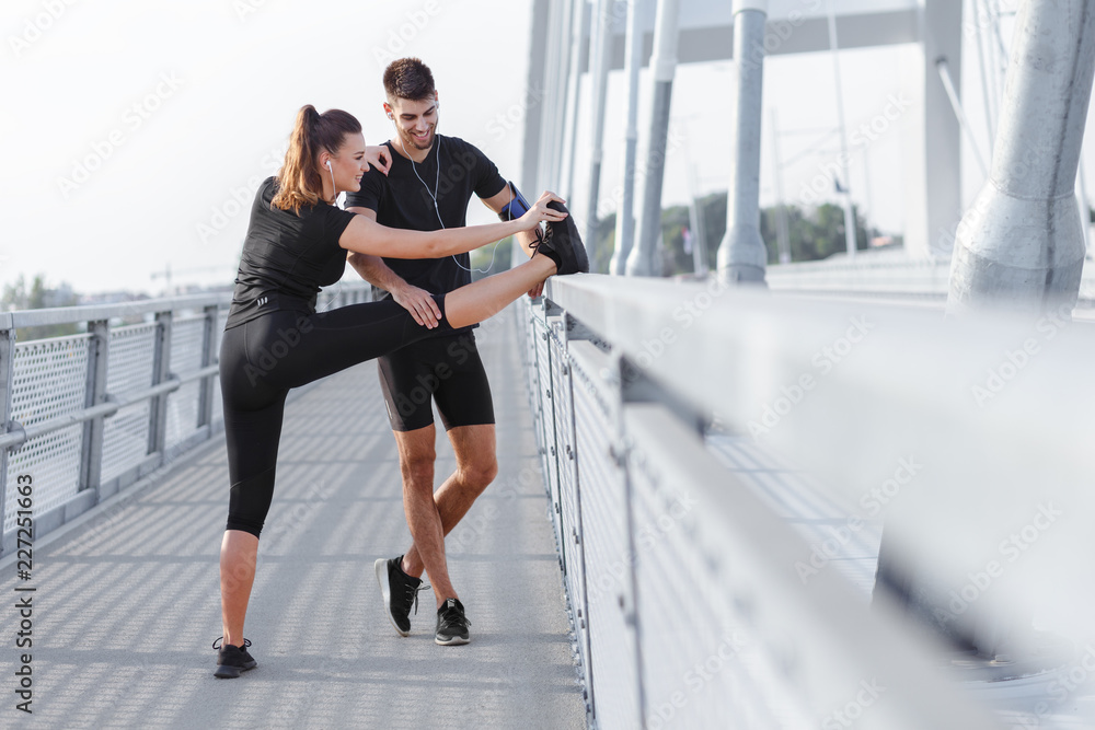 Obraz premium Young couple doing morning workout outdoors. Young man and woman stretching they muscle before running on bridge.