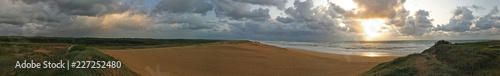 Panorama image of a beach or bay on the atlantic ocean with sand, grass, waves, high rocks and a blue sky with clouds on a sunny day during sunset