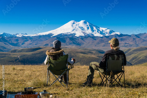 Man and woman sitting in the chairs admiring Elbrus mountain view landscape from the cliff edge. Enjoying nature vacation travel adventure at Caucasus mountains.