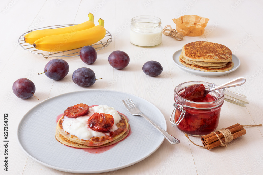 White wooden table with closeup image of dessert saucer with crumpets, pancakes, sour cream and fresh plums. The glass with cooked plums, fresh plums and fresh bananas in the background. White wooden table with closeup image of dessert saucer with crumpets, pancakes, sour cream and fresh plums. The glass with cooked plums, fresh plums and fresh bananas in the background.