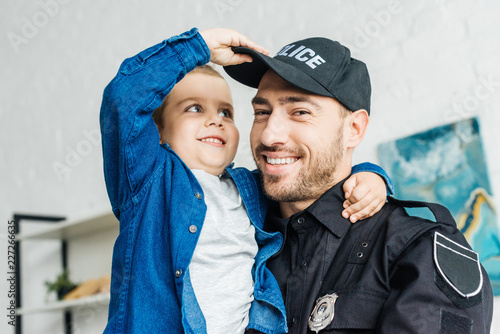 close-up portrait of smiling young father in police uniform carrying his little son and looking at camera photo