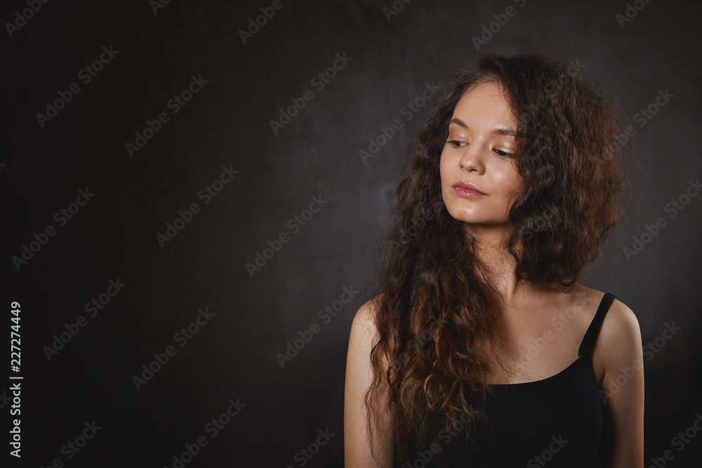 Astrology and mysticism concept. Gorgeous young female with voluminous long hair looking down and smiling mysteriously, posing against blank studio wall background with copyspace for your text