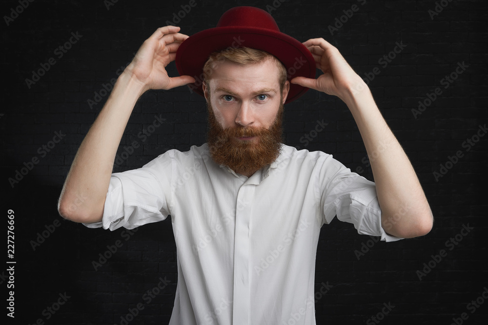 Attractive blue eyed young guy with ginger trimmed beard going to party, putting on stylish red round hat. Elegant young European male in white shirt, getting dressed, taking on trendy headwear