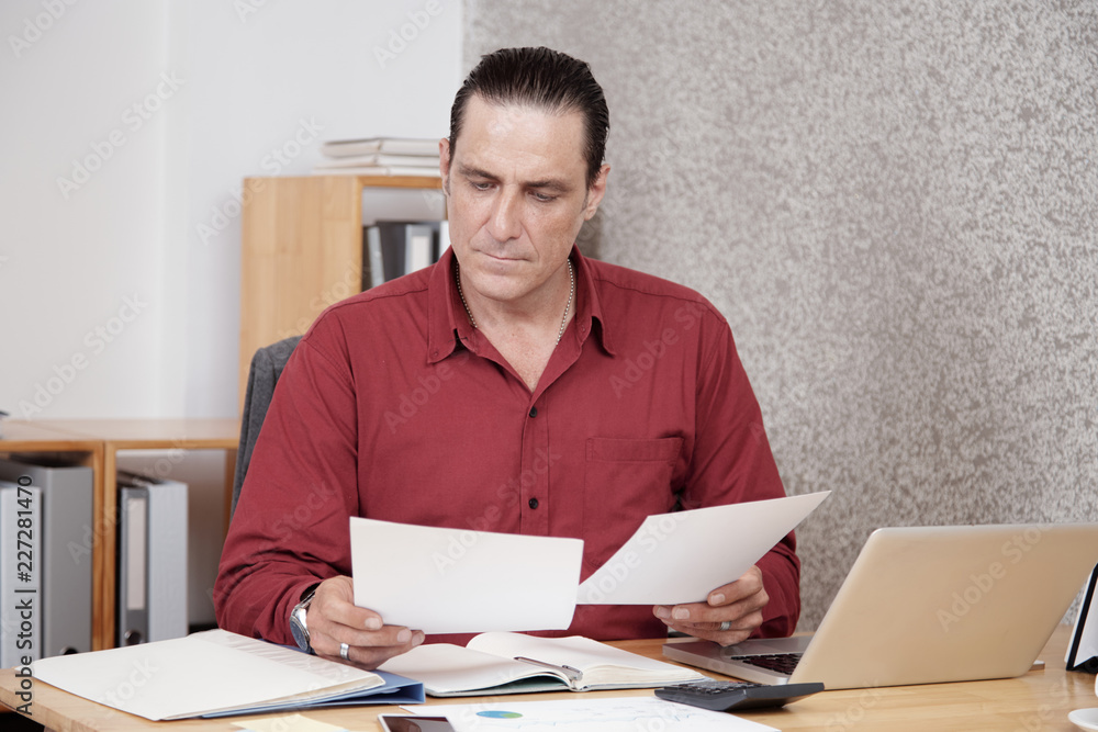 Serious office worker examining documents, he sitting at the table with laptop and calculator and doing financial report Serious office worker examining documents, he sitting at the table with laptop and calculator and doing financial report