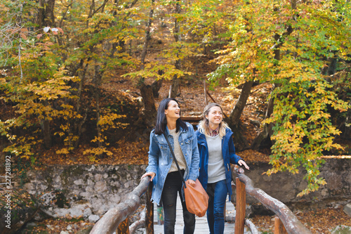 Two young girl friends walking on a wooden bridge in the autumn forest, talking and laughing.